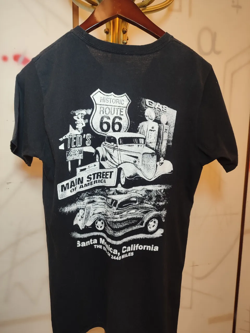 Remera negra de manga corta con estampa de la Ruta 66 y autos clásicos en la espalda. La estampa del frente es circular con el logo de la Ruta 66 y texto "Santa Monica, California The End of 2448 Miles". La estampa de la espalda muestra un paisaje con autos antiguos, una estación de servicio y el texto "Historic Route 66", "Ted's Main Street of America", "Santa Monica, California The End of 2448 Miles".Ancho hombros 0,42 Largo 0,72 ancho sisa 0,50