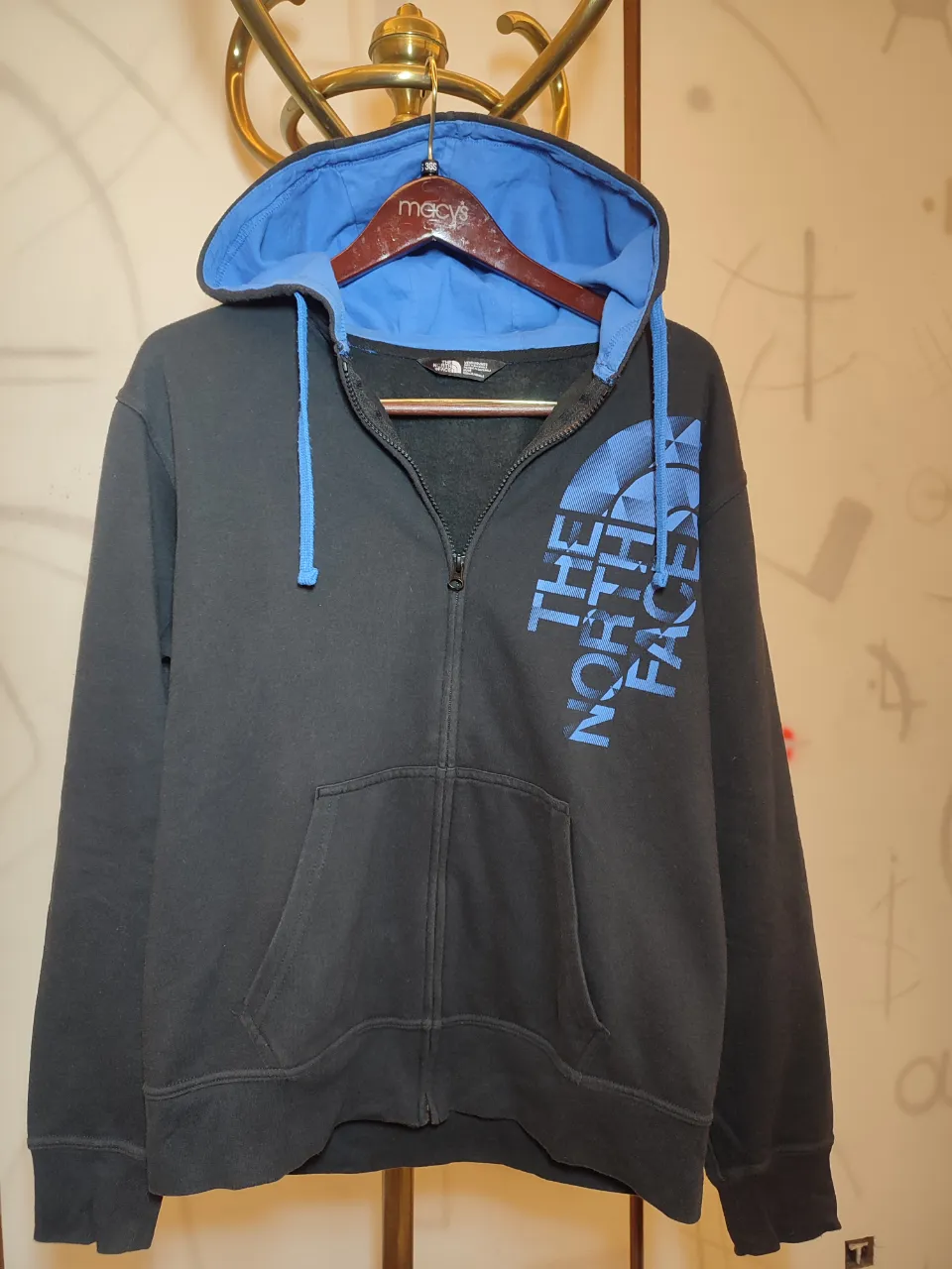 Buzo negro con capucha y cierre completo. Tiene cordones azules para ajustar la capucha y un logo grande de The North Face en azul en el frente. Posee bolsillos tipo canguro.largo de mangas 0,64.Ancho hombros 0,50.Lago 0,66.