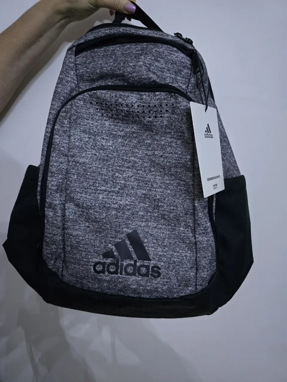 Mochila Adidas Defender Backpack, gris con detalles en negro. Ideal para llevar tus cosas al gimnasio o para el día a día. Cuenta con el logo de Adidas y etiqueta.