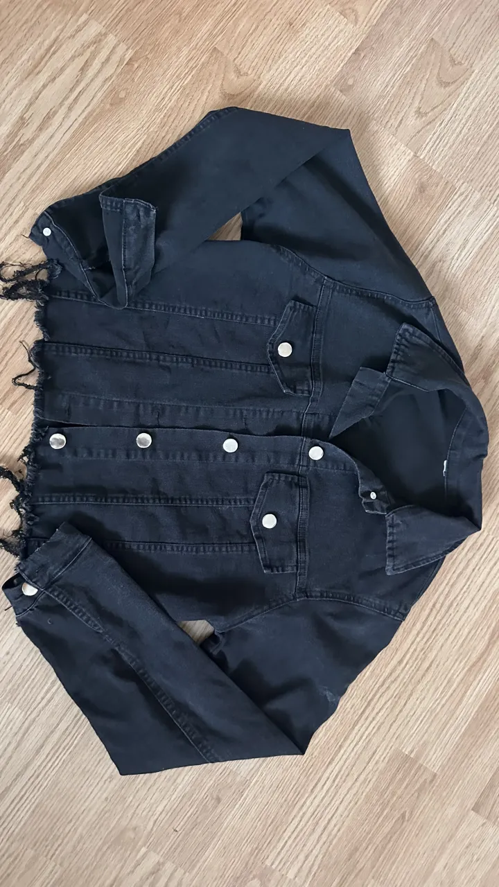 Campera de jean negra con botones plateados y detalles deshilachados en los puños y el ruedo. Posee bolsillos con tapa en el frente. Tiene un poco de spandex
