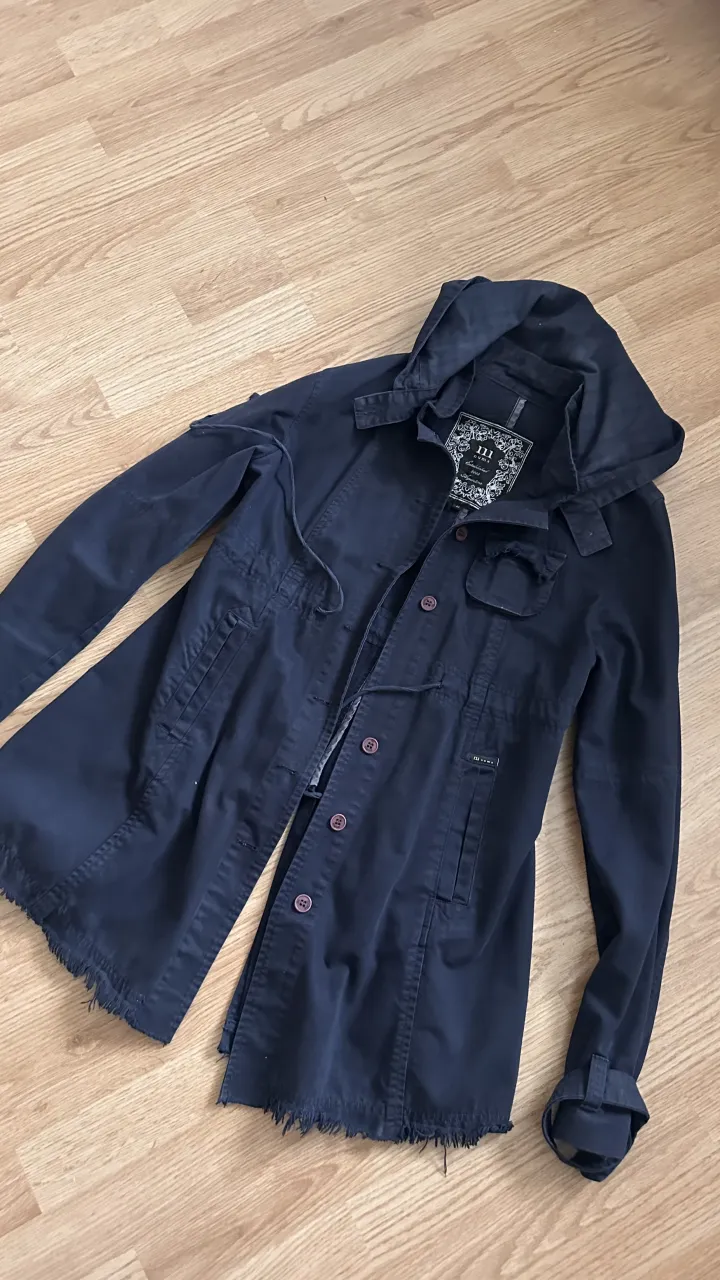 Campera azul marino con capucha, botones al frente y cordón de ajuste en la cintura. Posee bolsillos con solapa y puños con detalle de doblez. El ruedo es desflecado.