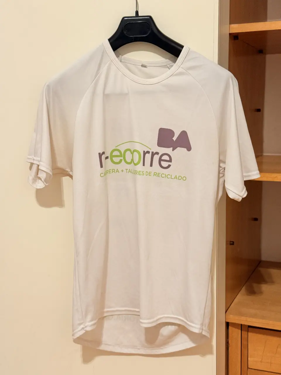 Remera deportiva blanca con logo de "r-ecorre" y "BA" en el frente. En la espalda, logos de "Buenos Aires Ciudad", "La Victoria" y otros sponsors. Ideal para actividades deportivas y eventos.