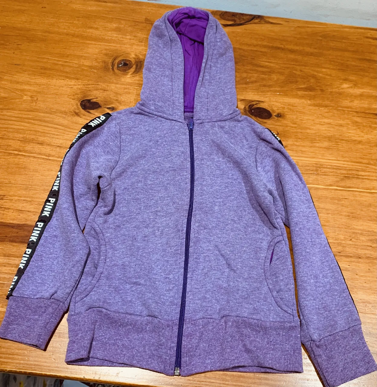Buzo con cierre y capucha para niña, color violeta. Con detalles de la marca PINK en la manga. Ideal para un look casual y cómodo.