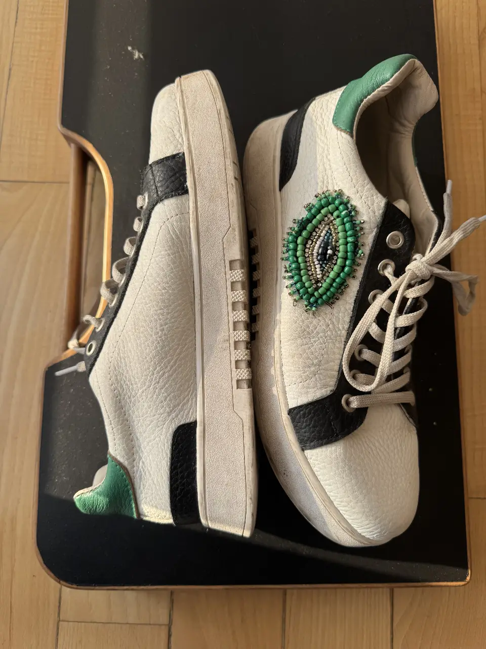 Zapatillas de cuero blancas con detalles en negro y verde, adornadas con un aplique de cuentas en forma de ojo. Suela gruesa y diseño moderno.