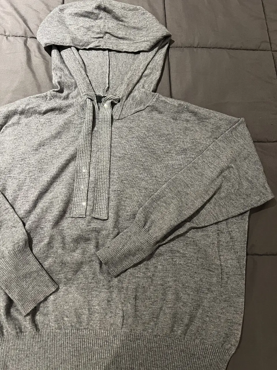 Buzo con capucha de color gris, con detalles de brillos en los cordones y puños tejidos. Ideal para un look casual y cómodo.