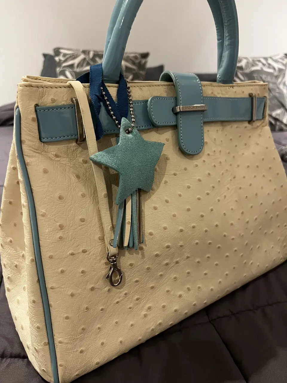 Cartera de mano beige con detalles en celeste, con textura tipo piel de avestruz y dije de estrella. Ideal para un look elegante y sofisticado.