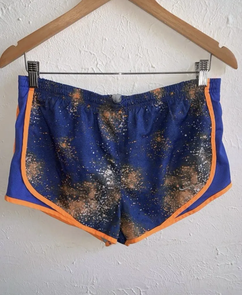 Shorts deportivos Nike Dri-Fit azules con estampado tipo tie-dye en tonos naranja y blanco. Tienen vivos naranjas en los laterales y el logo de Nike bordado en plateado.