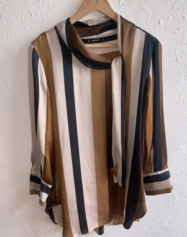 Blusa de seda con estampado de rayas verticales en tonos marrones, beige, negro y blanco. Posee cuello con lazo y puños anchos.