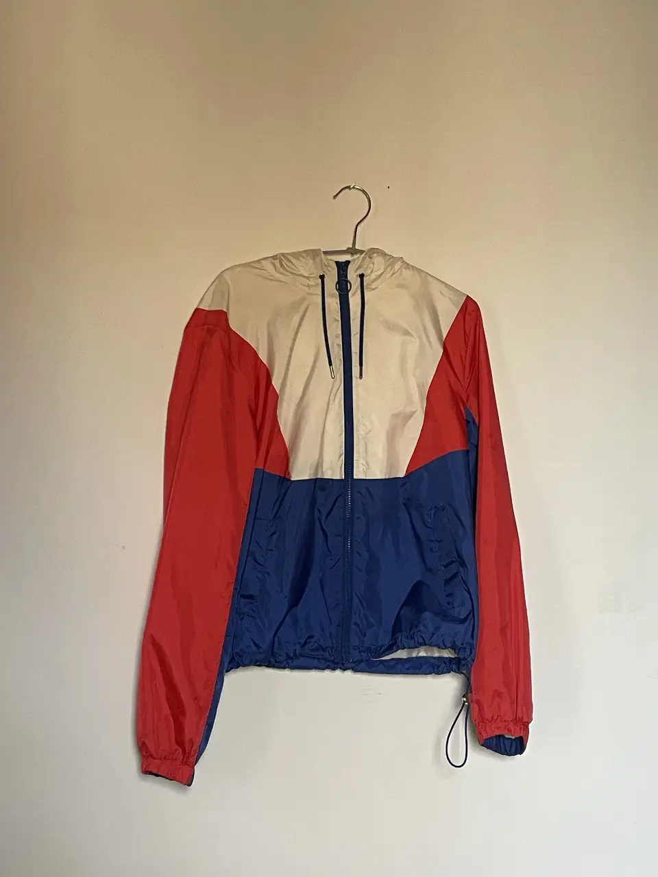 Rompevientos deportivo con diseño tricolor en rojo, blanco y azul. Posee capucha y cierre frontal. Ideal para actividades al aire libre.
