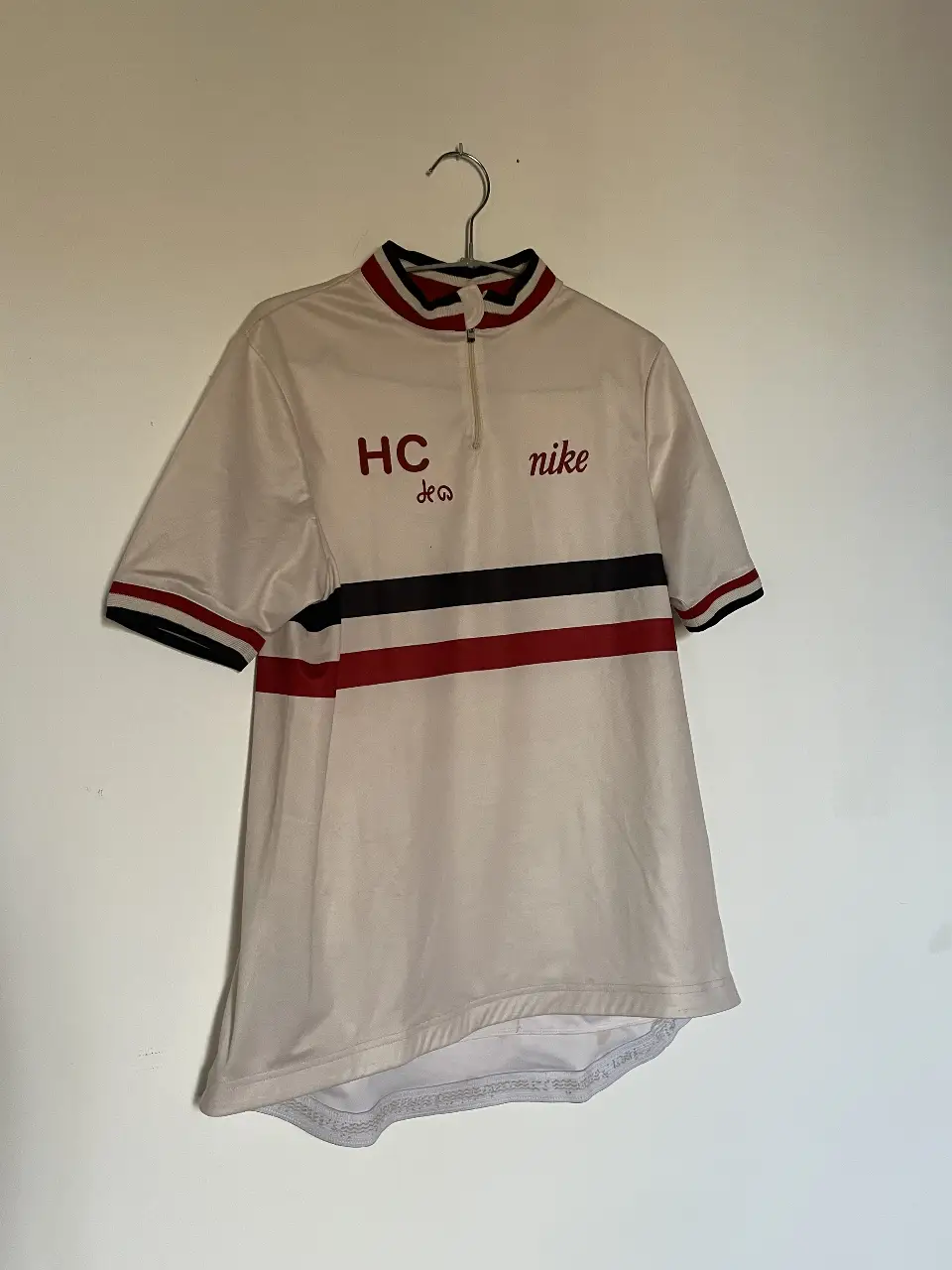 Camiseta de ciclismo Nike vintage, color crema con detalles en rojo, negro y azul. Con logo "HC" y "nike" estampado. Ideal para los amantes del ciclismo retro.