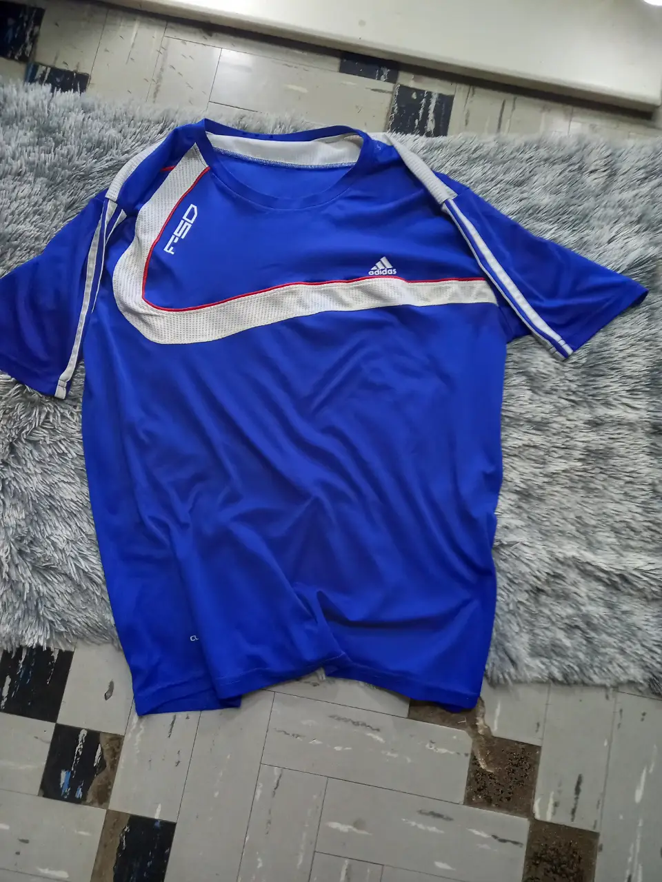 Remera deportiva Adidas azul con detalles blancos y rojos, ideal para entrenamientos o uso casual. Diseño con logo de la marca y detalles en las mangas.tiene en la axila un contorno que no se llega a casi notar pero si te acercas se puede ver, de igual manera casi no se nota 