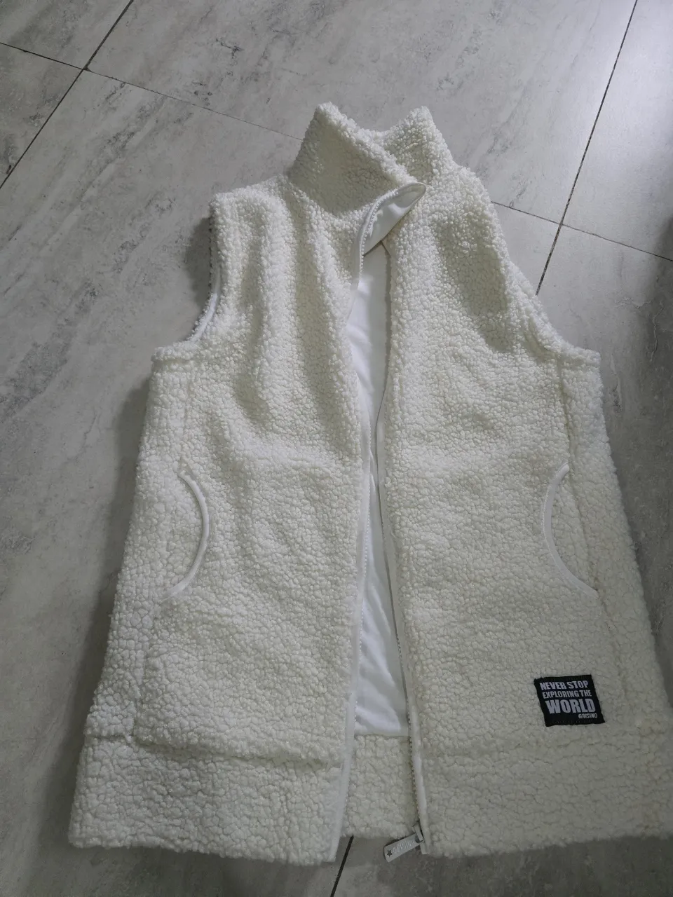 Chaleco blanco de peluche con cierre y bolsillos laterales. Tiene un parche con texto en la parte inferior derecha. Super abrigado. Es como un cs de adultos xq lo compré para mi q mido 1.54 y peso 55 kg.