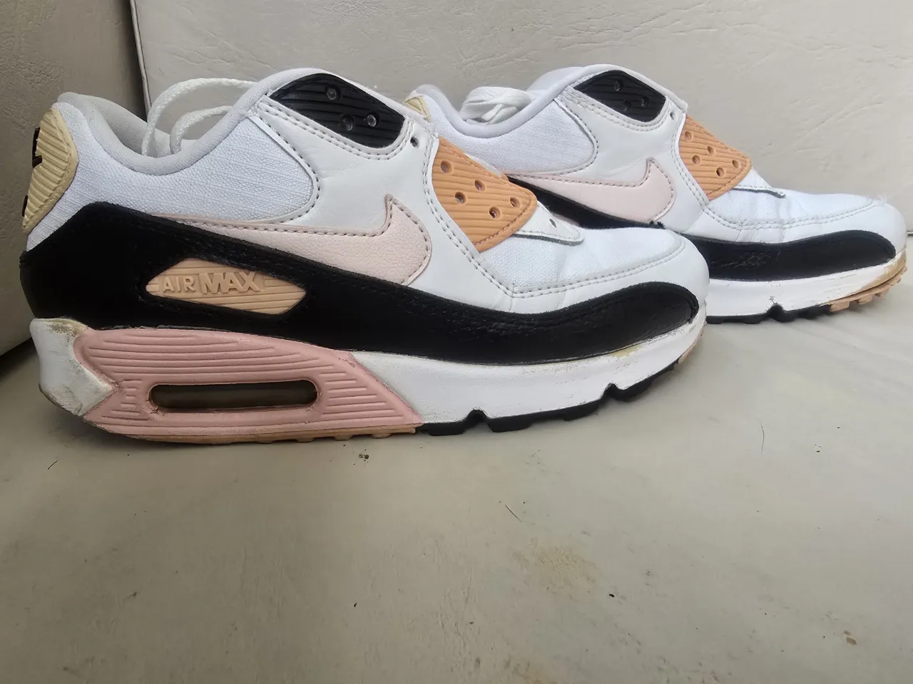 Zapatillas deportivas blancas con detalles en negro, rosa pálido y naranja. Presentan el logo de Nike y la inscripción "Air Max" en los laterales.