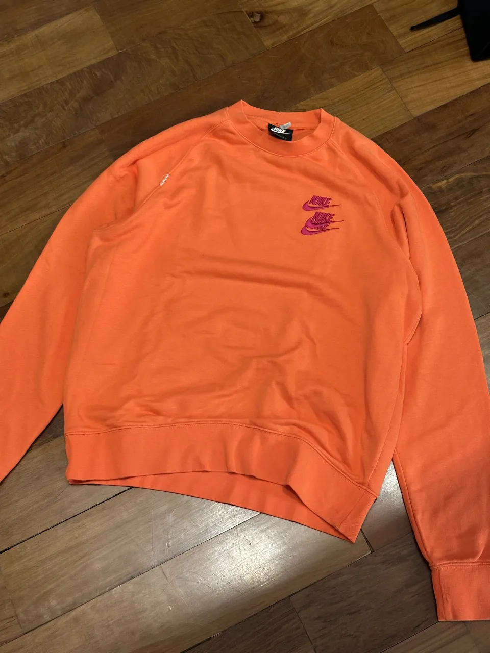 Buzo naranja con doble logo de Nike bordado en color fucsia en el frente. En la espalda, tiene un estampado con nombres de ciudades en color fucsia: Tokyo, Barcelona, London, Shanghai, Paris, Milan, Los Angeles, New York, Beijing, Mexico City, Berlin, Seoul. En la nuca, tiene bordado "Beaverton, Oregon" en color verde claro.