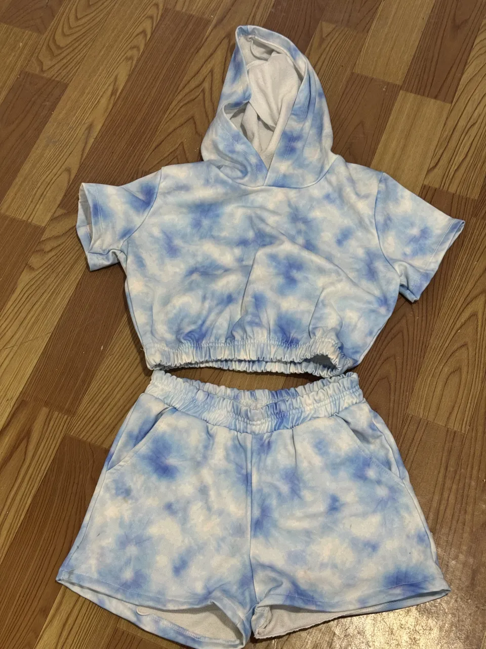 Conjunto de short y buzo crop con capucha, ambos con estampa tie-dye en tonos de azul y blanco. El buzo tiene mangas cortas y elástico en la cintura. El short tiene cintura elastizada y bolsillos.