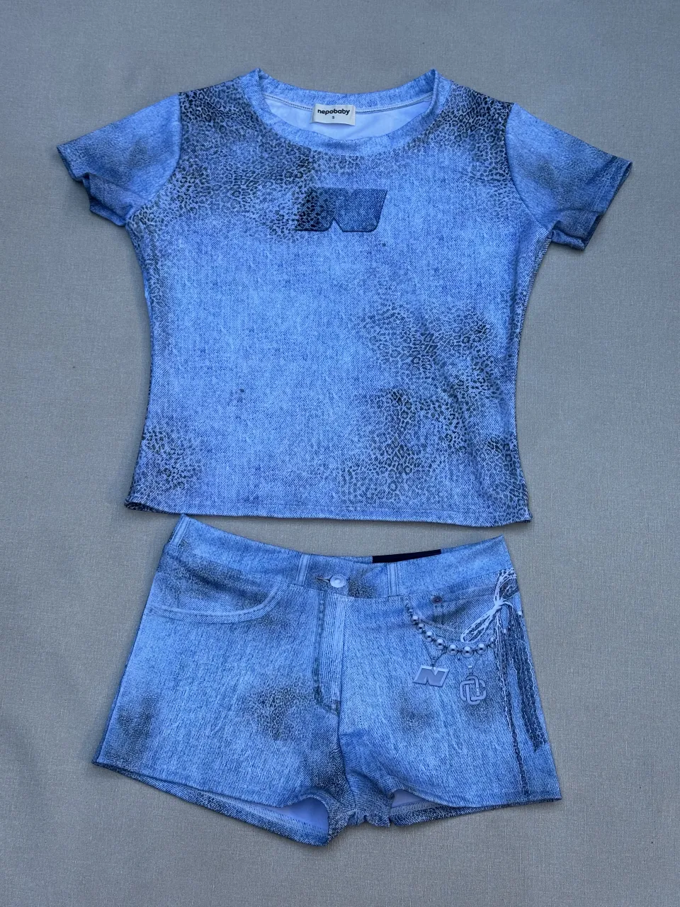 Conjunto de remera y shorts con estampa efecto "jean" y detalles de cadenas y dijes. La remera es de manga corta y cuello redondo. Los shorts son tiro alto con bolsillos.