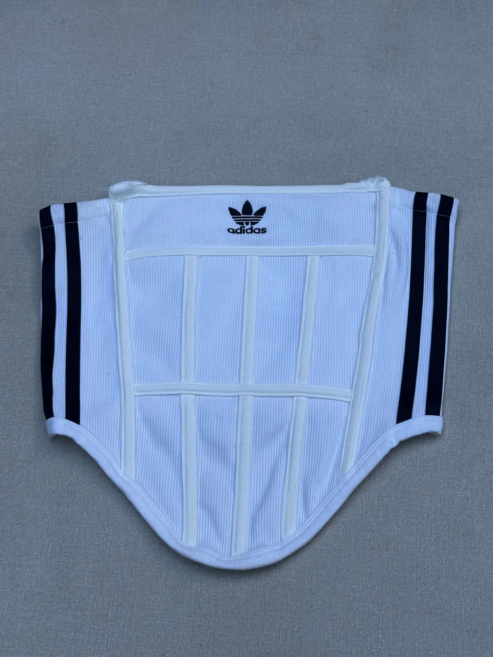 Top corset blanco de Adidas con las icónicas tres tiras negras a los costados. Detalle: Arriba uno de los palitos se sale por que esta roto. 