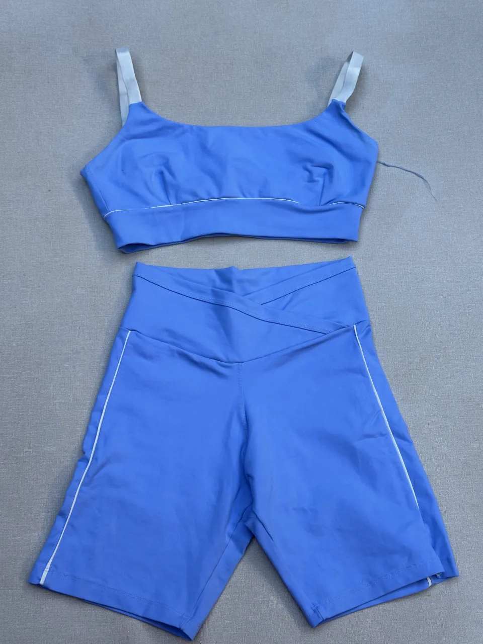 Conjunto deportivo de top y shorts de ciclista en un vibrante color azul. El top cuenta con tirantes blancos y detalles de ribete plateado, mientras que los shorts presentan un diseño cruzado en la cintura y también tienen ribetes plateados a los costados. Ideal para entrenamientos o para un look casual y cómodo.