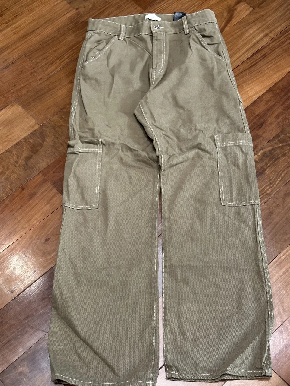 Pantalón cargo de color verde militar con costuras blancas visibles. Posee bolsillos frontales y dos bolsillos tipo parche en los laterales de los muslos. Cierre con botón y cierre.