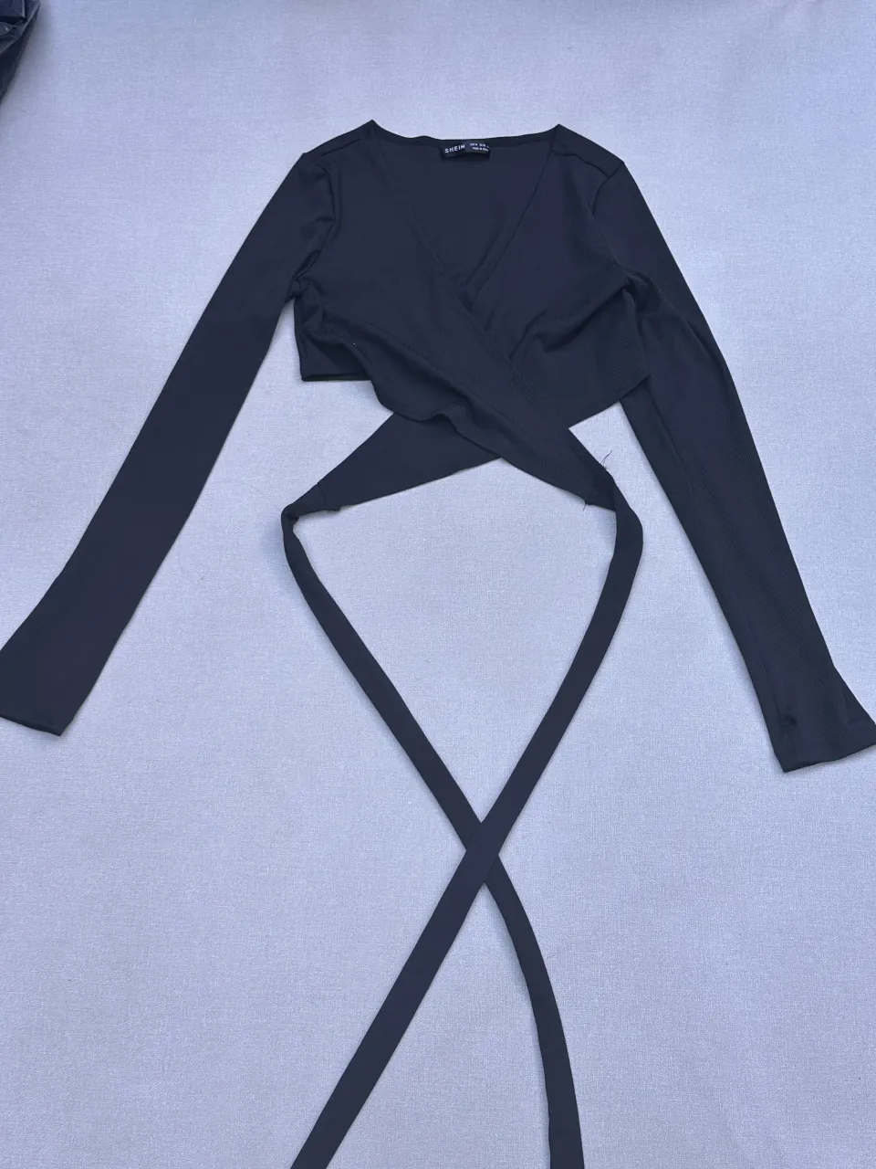 Top negro de Shein con mangas largas y escote cruzado. Posee tiras para atar en la cintura. Tiene un detalle de rotura en una de las mangas para que quede tipo guante