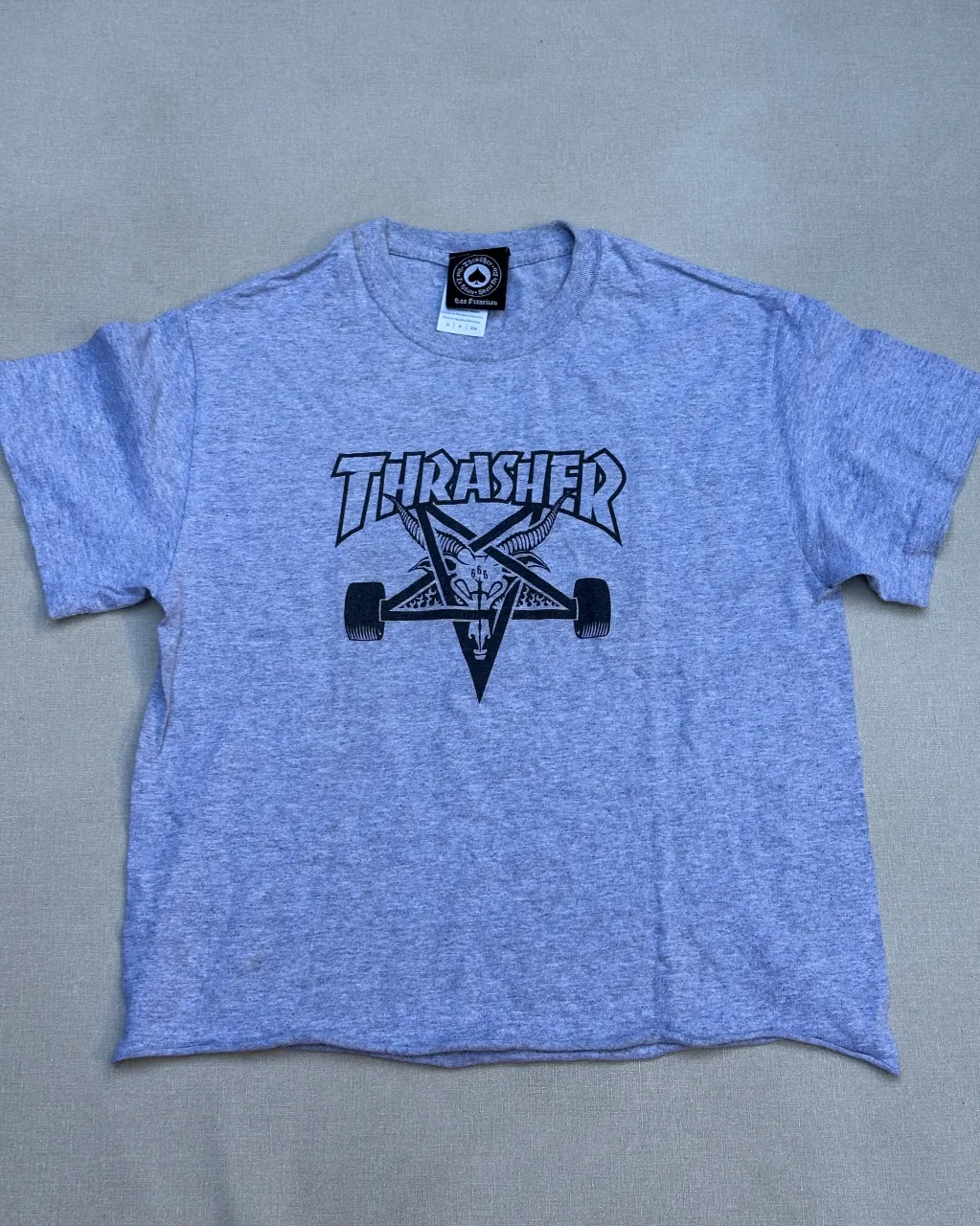 Remera gris de manga corta con estampa de "Thrasher" y un pentagrama con cabeza de cabra en el frente. Ideal para un look urbano y canchero.