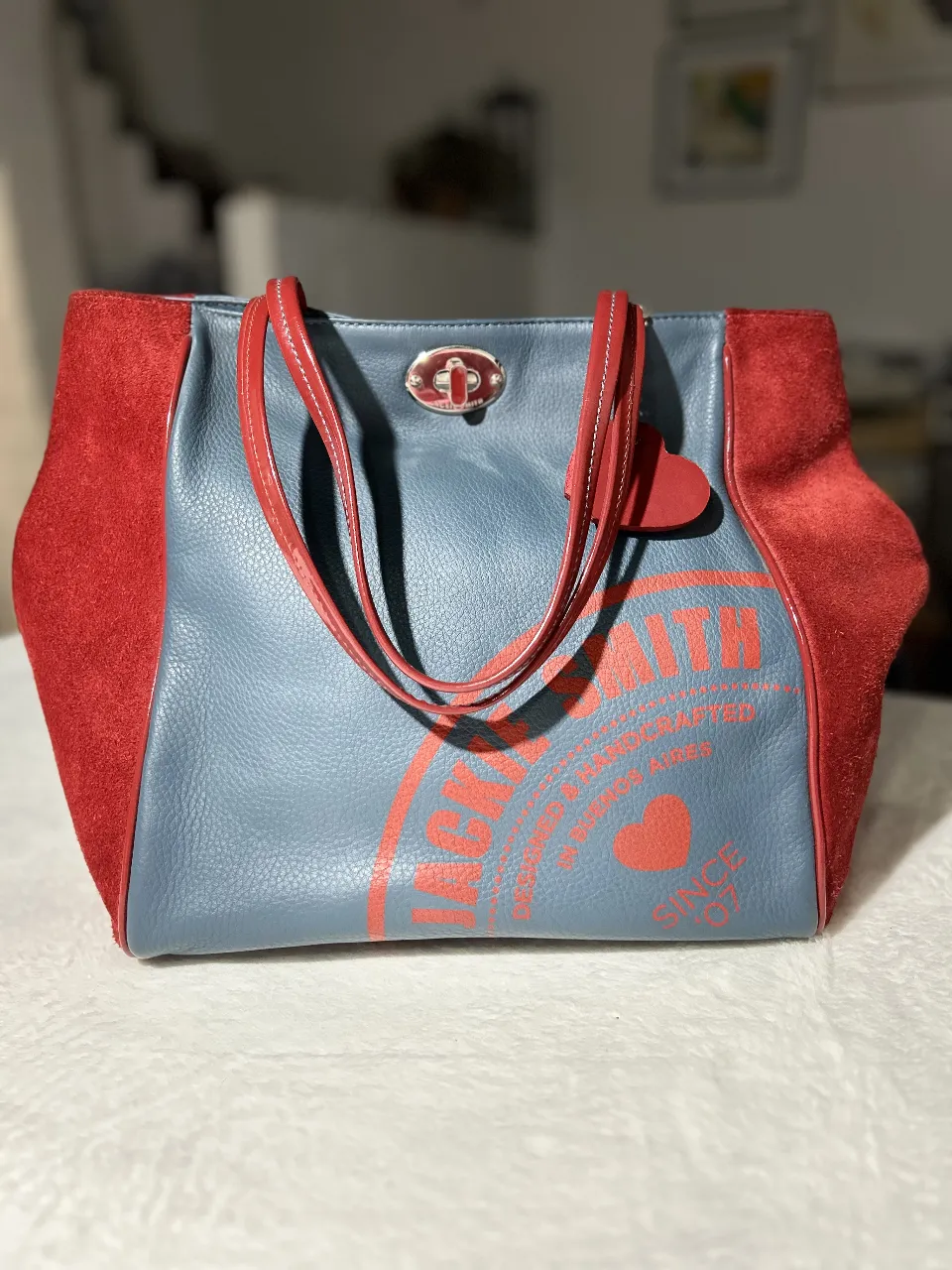 Cartera tipo tote bag de cuero sintético en color azul con detalles en gamuza roja en los laterales. Posee manijas rojas y un cierre con un detalle metálico. En el frente, tiene un estampado en rojo. Ideal para un look casual y moderno.