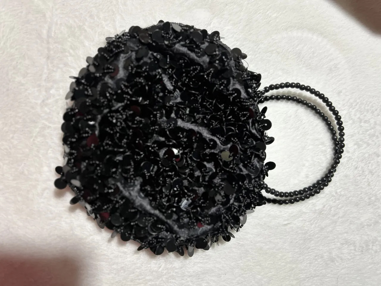 Mini cartera de mano redonda, adornada con lentejuelas negras y un asa de cuentas a juego. Ideal para eventos especiales o para darle un toque glamoroso a cualquier look.