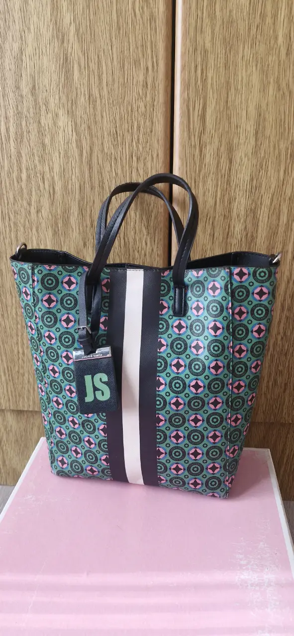 Bolso tote con estampado geométrico en tonos verde y rosa. Detalles en negro y una franja blanca central. Incluye un llavero con las iniciales "JS". Ideal para un look casual y moderno.