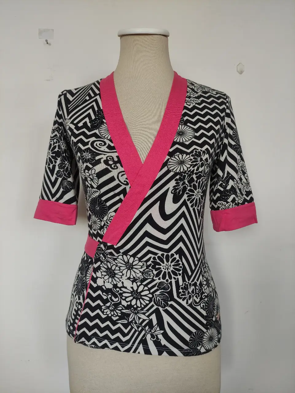 Kimono con estampado geométrico en blanco y negro, con detalles en rosa. Diseño cruzado con mangas 3/4 y cinturón a juego. Ideal para un look elegante y moderno.