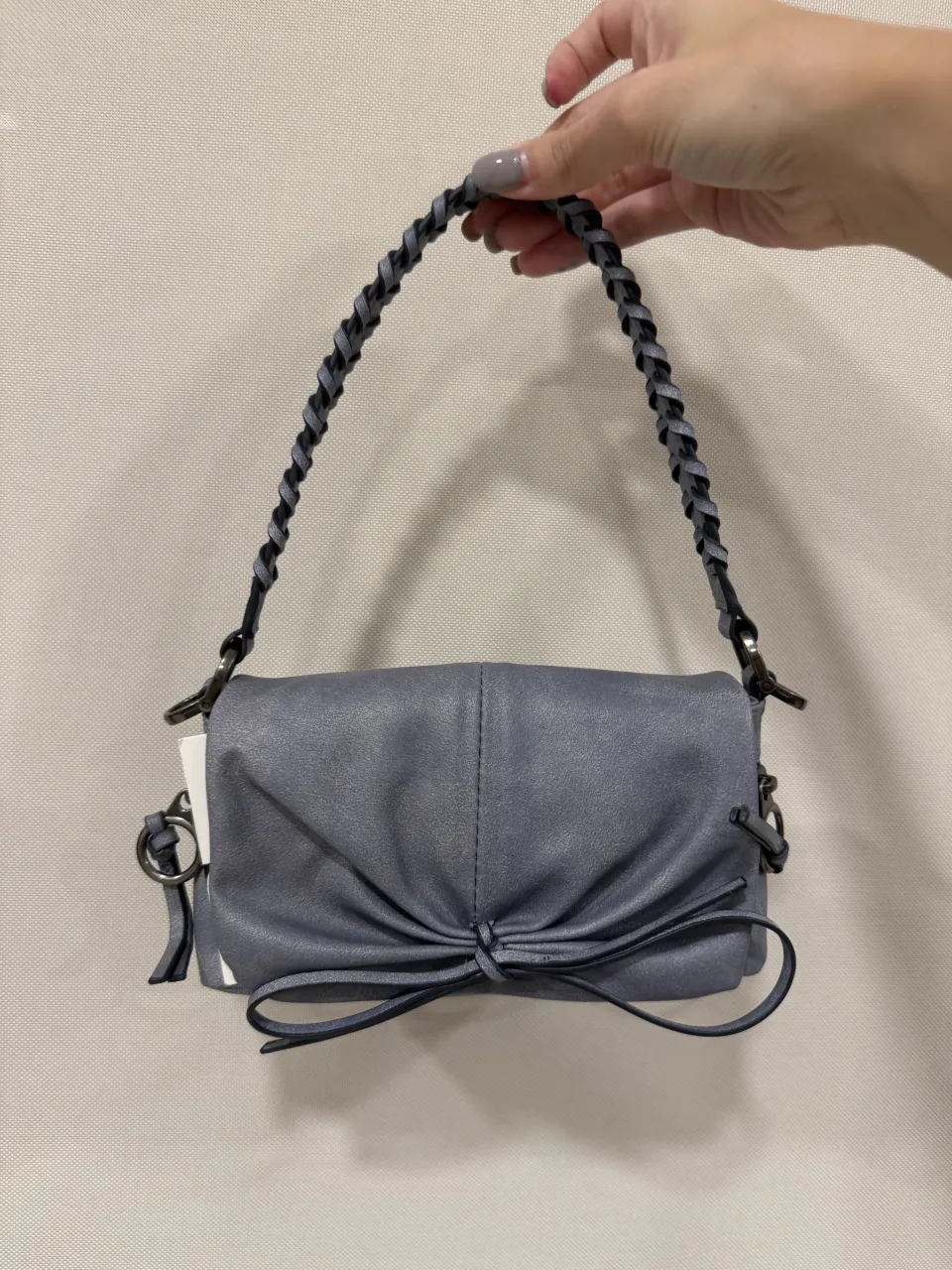 Cartera de mano color gris perla, con manija trenzada y detalle de moño en la parte frontal. Posee dos bolsillos con cierre en la parte delantera y uno en la parte trasera. Ideal para complementar looks casuales y de fiesta.