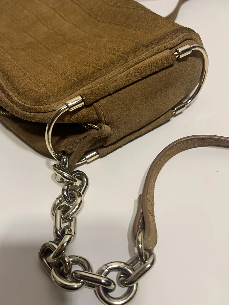 Cartera bandolera de gamuza marrón con detalles plateados y cadena. Ideal para un look casual y moderno.