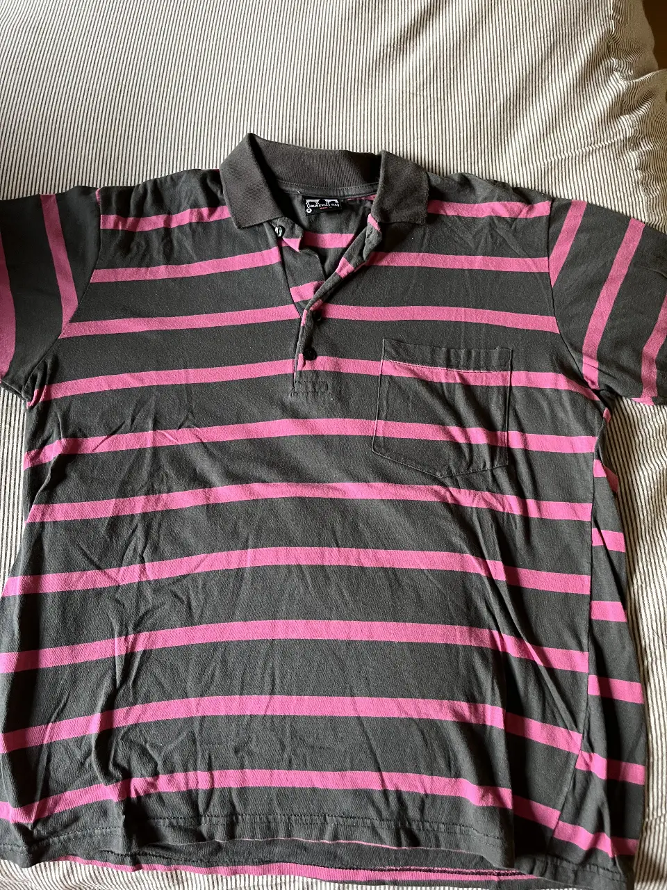 Remera polo a rayas horizontales en gris y rosa, con cuello y bolsillo. Ideal para un look casual y cómodo.