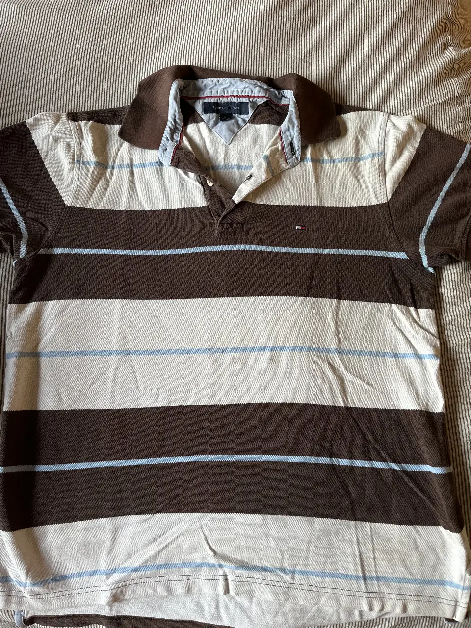 Remera polo Tommy Hilfiger a rayas horizontales en tonos marrón y blanco. Cuello y mangas en marrón oscuro. Logo de la marca bordado en el pecho. Talle M.