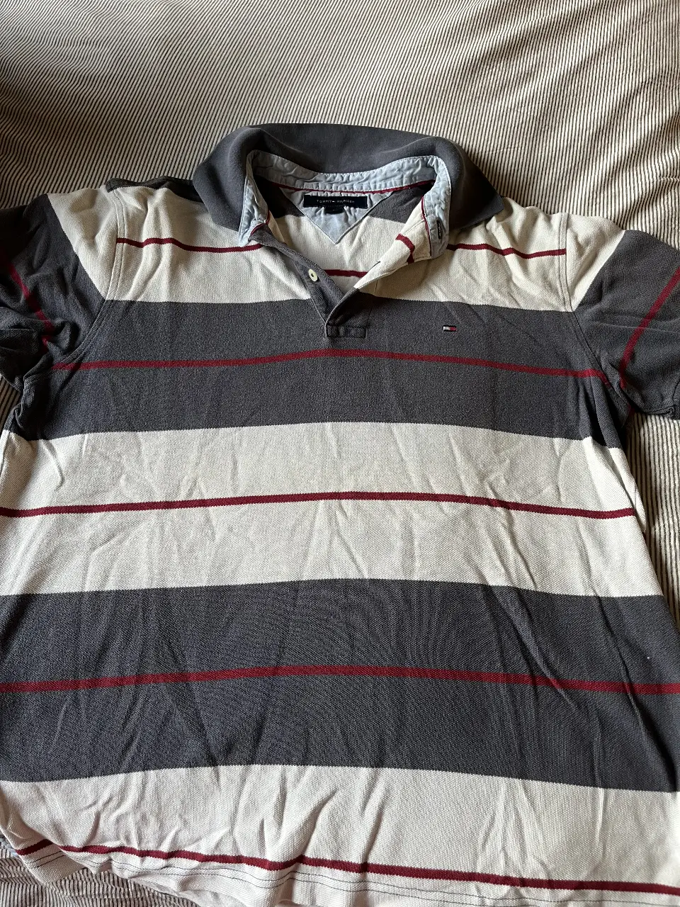 Remera polo a rayas horizontales en tonos grises y blanco, con detalles en rojo. Cuello y mangas en gris oscuro. Logo Tommy Hilfiger bordado. Talle M.
