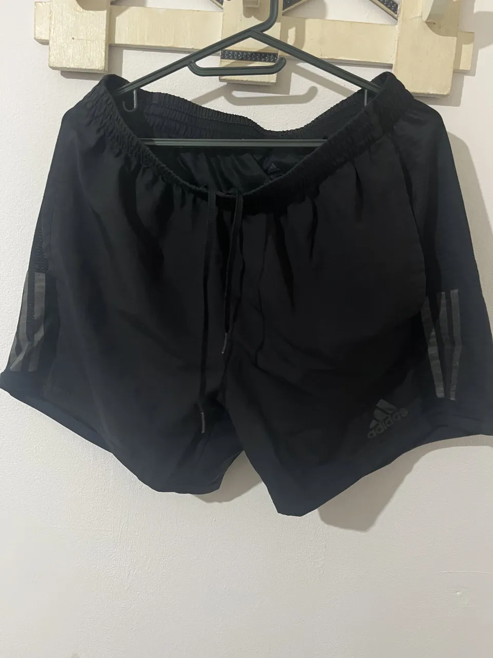 Shorts deportivos negros con tres franjas grises reflectivas en el lateral. Cintura elastizada con cordón de ajuste. Logo de Adidas y "AEROREADY" estampado.