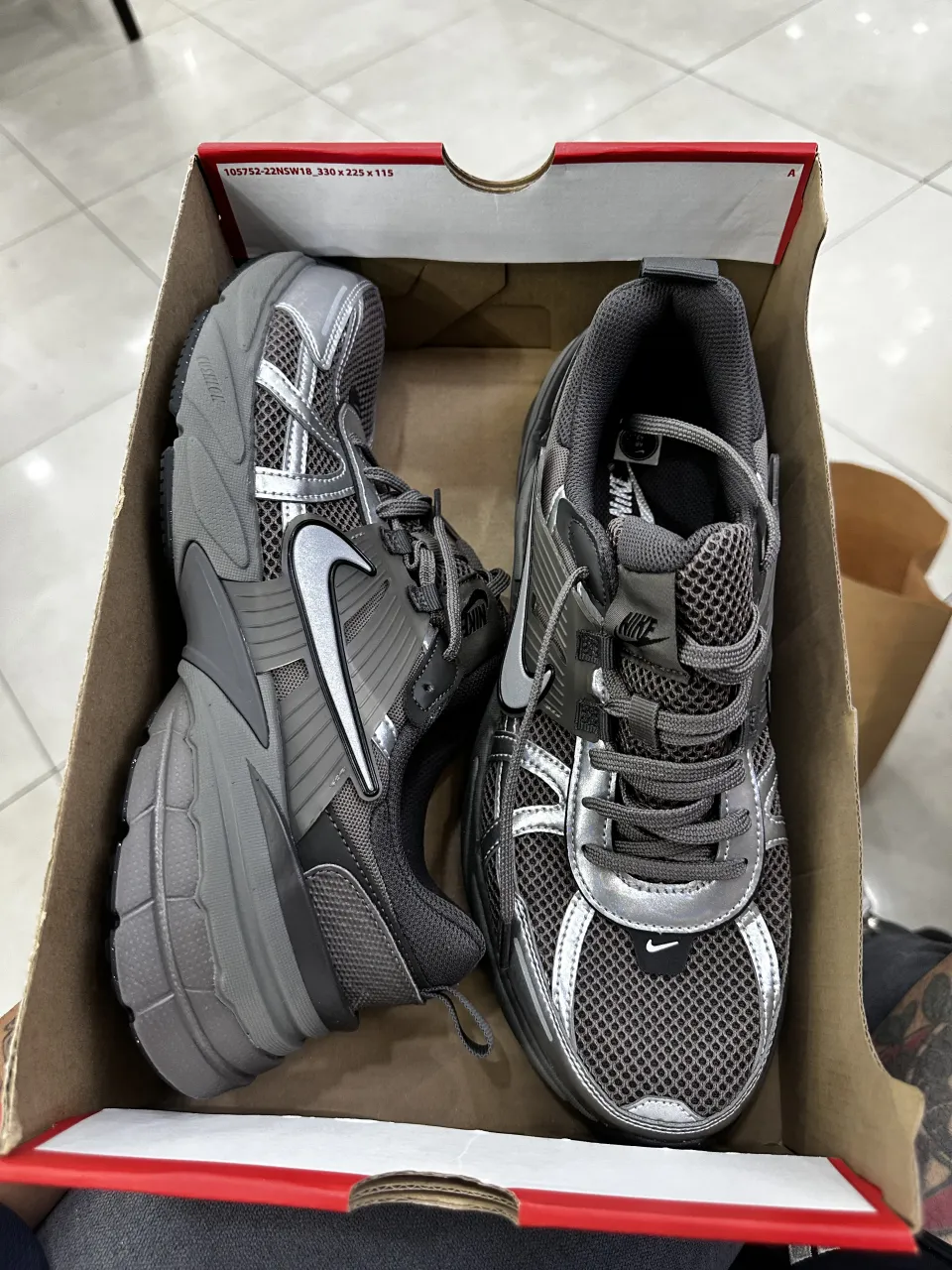 Zapatillas deportivas Nike V2K Run en color gris y plateado. Presentan el logo de Nike en los laterales y en la lengüeta. La suela es gruesa y de goma.