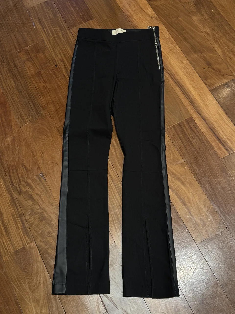 Pantalón negro tiro alto con detalles de cuero en los laterales y cierre metálico. Posee tajos en los bajos de las piernas.