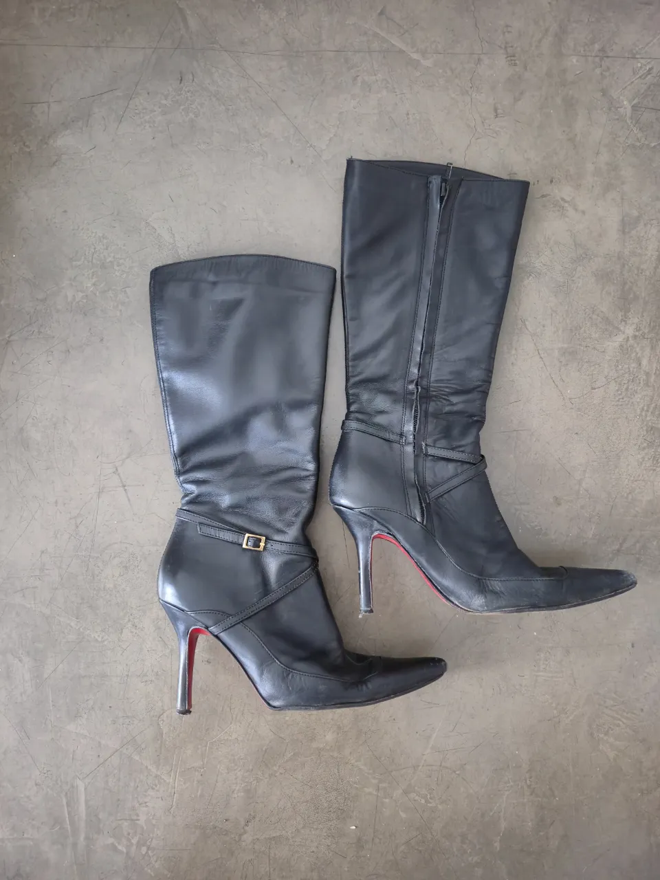 Botas de cuero negras, caña alta, con taco aguja y punta fina. Detalle de hebilla dorada en el lateral de una de las botas. Suela roja.