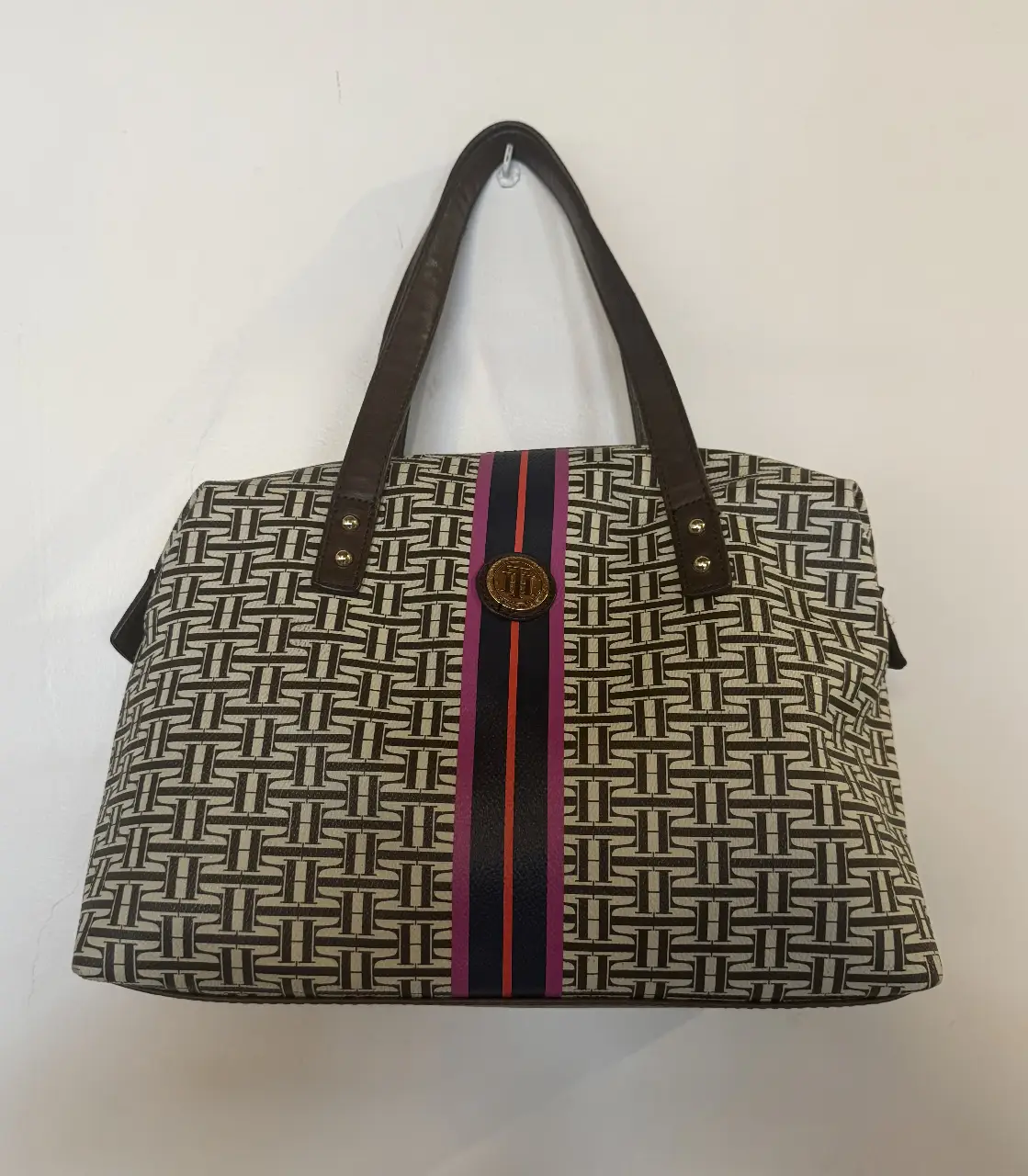 Cartera tipo tote bag de mujer, con estampado geométrico en tonos marrones y detalles en negro, con una franja central en colores vibrantes. Posee manijas marrones y un aplique dorado con el logo de la marca. Ideal para llevar tus pertenencias con estilo y organización.