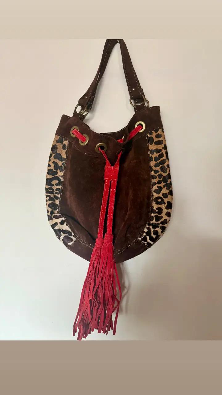 Cartera tipo hobo de gamuza marrón con detalles de estampado animal print en los laterales. Cuenta con un cierre de cordón rojo y flecos a juego. Ideal para un look bohemio y con estilo.