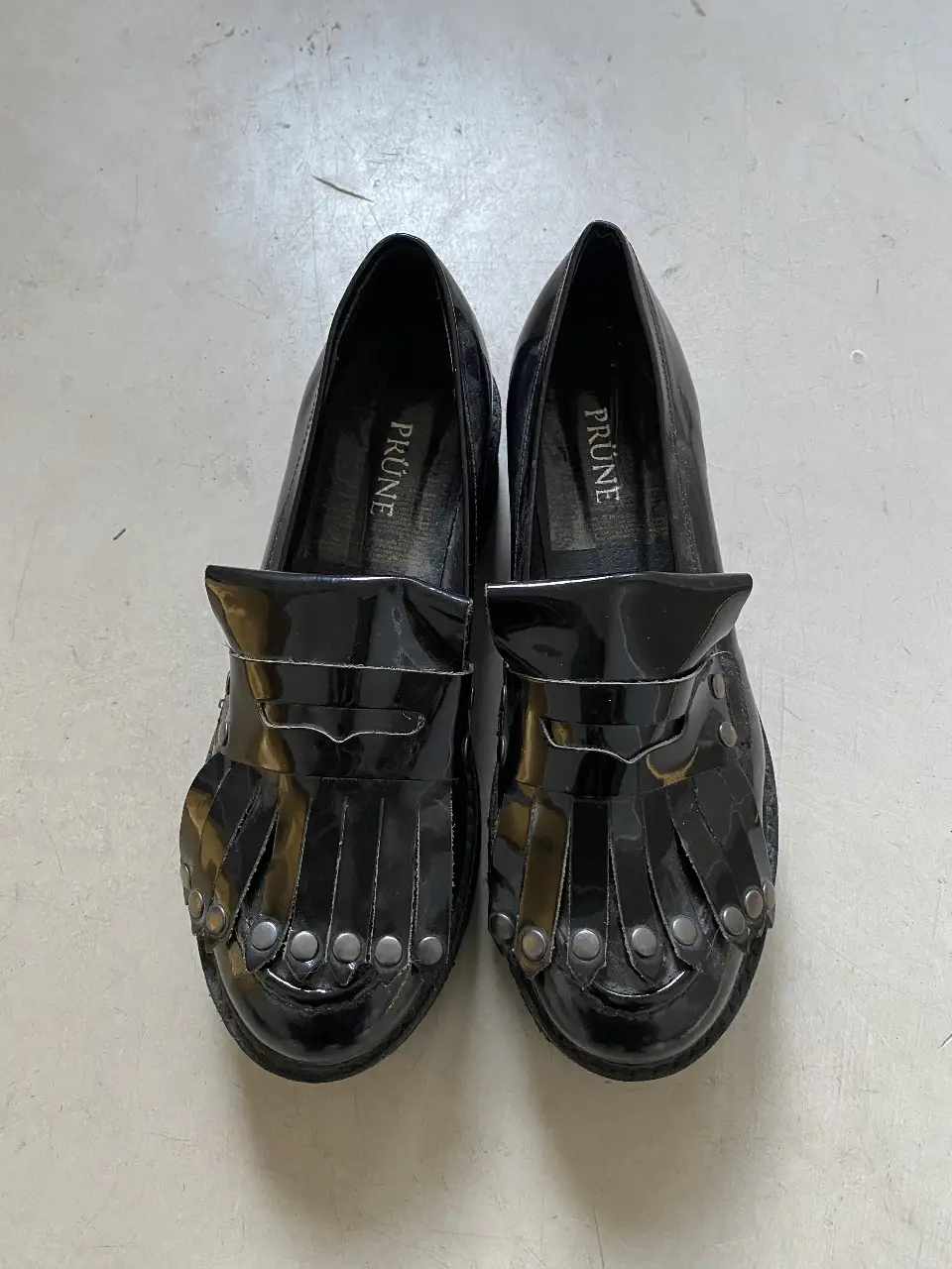 Mocasines PRUNE negros de charol con detalles de tachas y flecos. Ideales para un look elegante y moderno. 