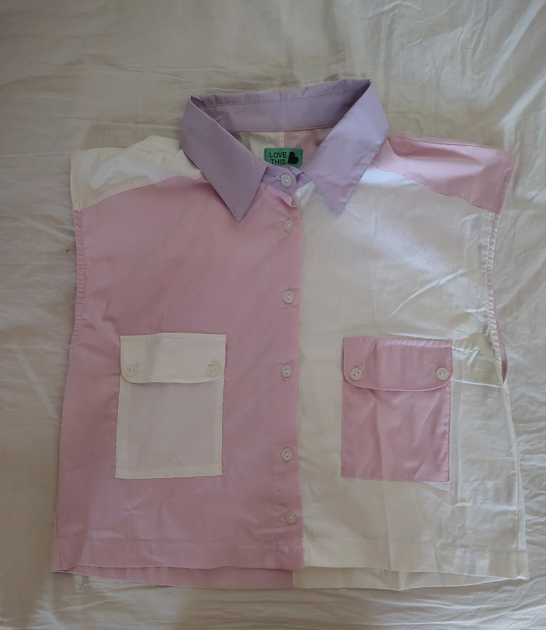 Remera sin mangas con cuello de camisa, diseño color block en blanco y rosa pastel. Posee dos bolsillos frontales con solapa y botones. Ideal para un look casual y fresco.