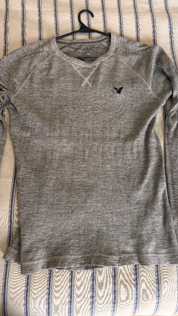 Sweater de hombre American Eagle, manga larga, color gris, con logo bordado en el pecho. Ideal para un look casual y cómodo. 2x1
