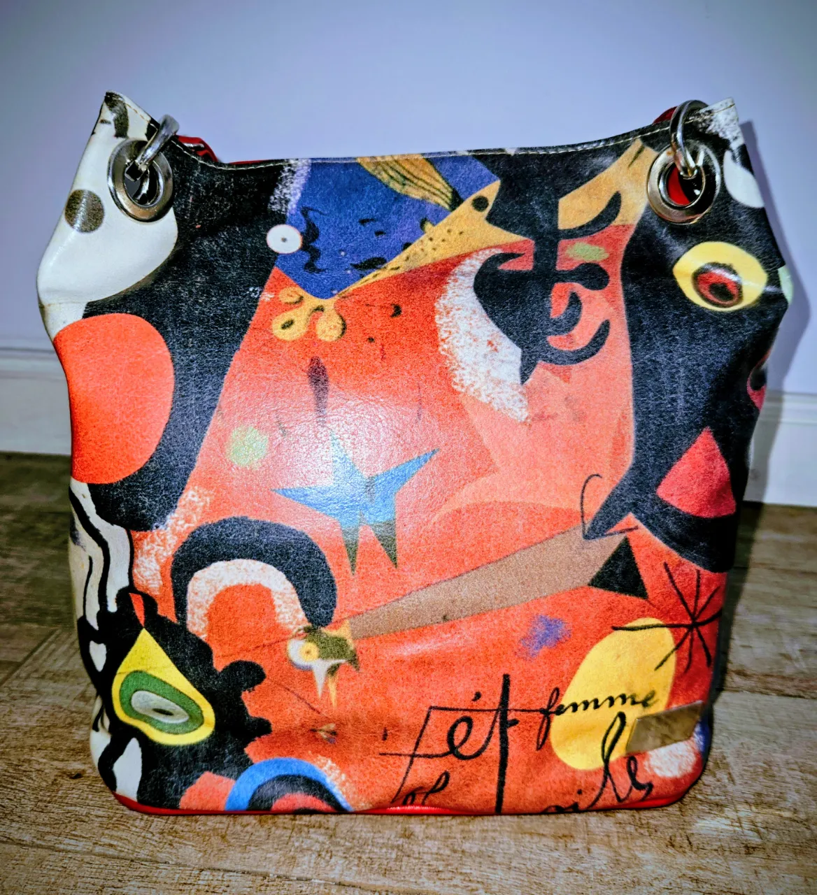 Bolso de cuero con un estampado artístico vibrante y abstracto, lleno de colores y formas que recuerdan a la obra de Miró. Tiene un diseño único y llamativo, ideal para darle un toque original a cualquier outfit. Posee dos argollas metálicas en la parte superior para sujetar las asas y cierre.