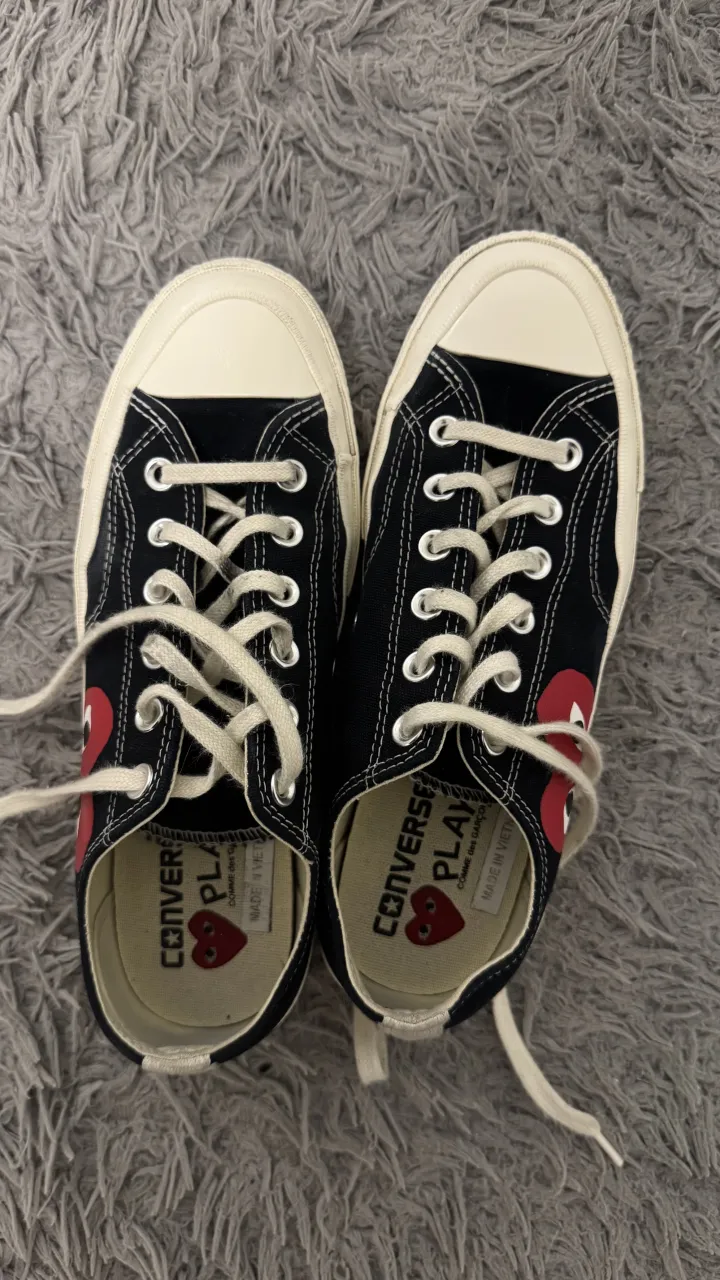 Zapatillas bajas de lona negras con detalles rojos en forma de corazón y cordones blancos. La suela es de goma color marrón.