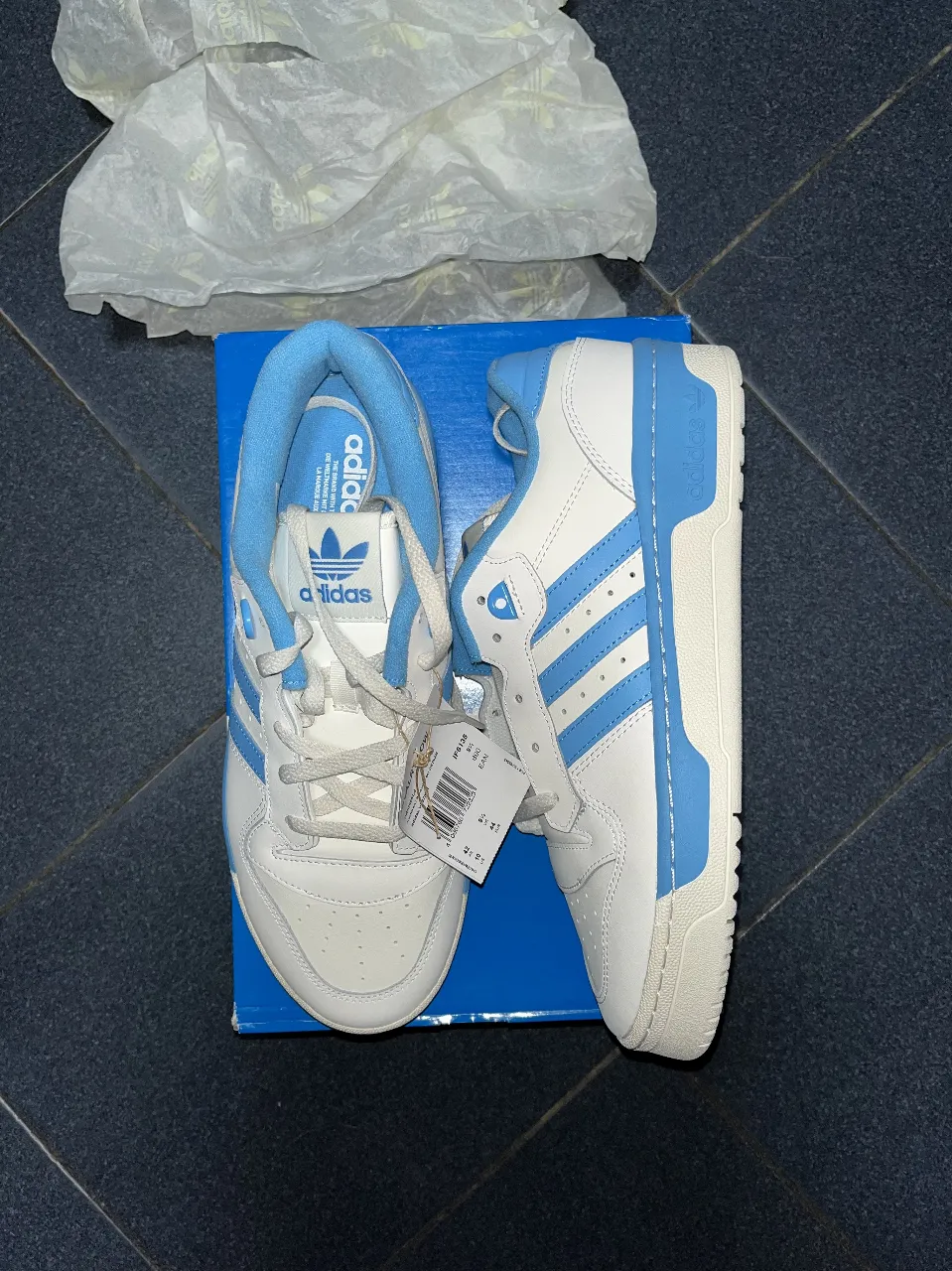 Zapatillas Adidas Rivalry Low en color blanco con detalles en celeste. Diseño clásico y urbano, ideales para un look casual y cómodo.