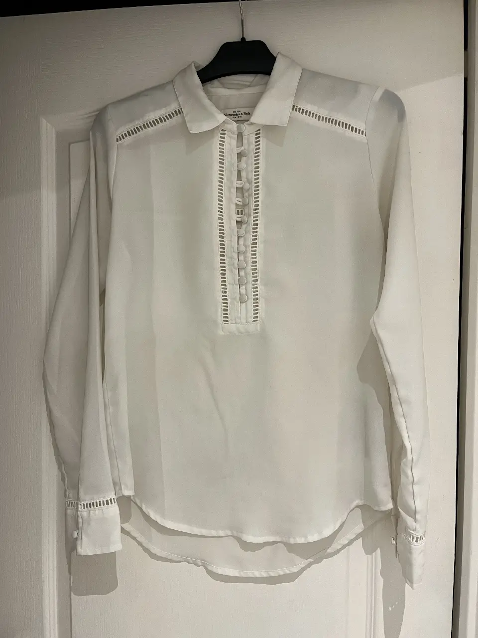 Blusa blanca de Abercrombie & Fitch, con detalles calados en hombros y pecho. Cuello camisero y cierre con botones. Ideal para un look elegante y versátil.