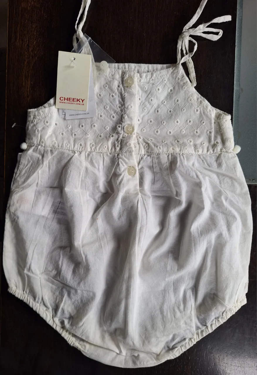 Enterito blanco de algodón para bebé, con delicados bordados en la parte superior y detalles de pompones. Posee botones en la parte trasera y tiras regulables en los hombros.