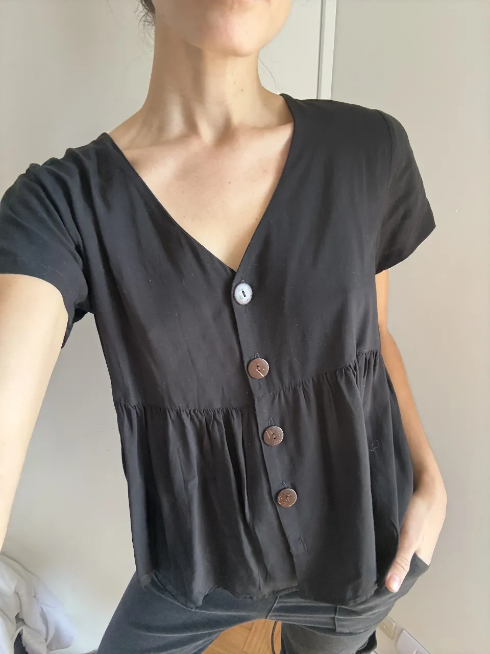 Remera negra con escote en V y botones de coco. Tiene un corte babydoll con frunces en la cintura que le da un movimiento suelto y canchero. Ideal para un look casual y cómodo.