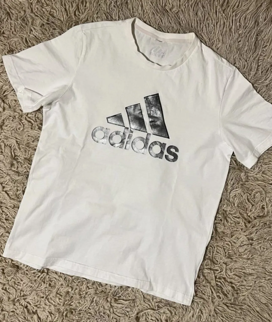 Remera blanca de manga corta con logo grande de Adidas estampado en el frente. El logo es de color gris metalizado con líneas finas. etiqueta interior cortada. Sisa 53 Cm - Largo 71 Cm 


